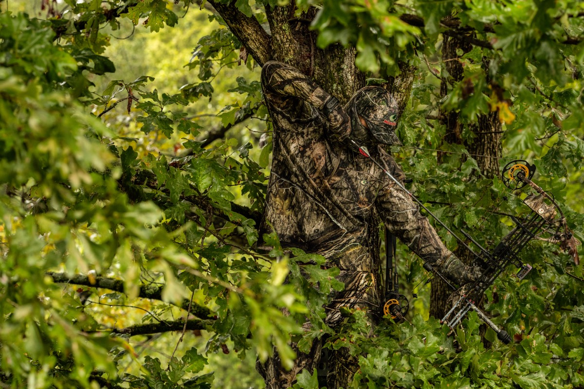 man concealed with camo draws back bow in a tree